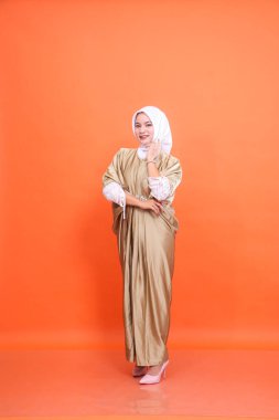 Kaftan başörtüsü takan, kolları çenesinde turuncu bir arka planı olan gülümseyen Asyalı Müslüman bir kız. İslam dini, moda ve ticari konsepti