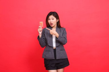 Asyalı iş kadınının ifadesi kameraya şok oldu, elinde bankamatik kartı vardı, kırmızı arka planda ceket ve etek giyiyordu. Finansal, iş ve reklam kavramları için