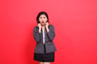 Endonezyalı ofis kadınının ifadesi şoke olmuş, iki eli göğsünün önünde birbirine kenetlenmiş, gri bir ceket ve kırmızı arka planda mini etek giymiş. moda, yaşam tarzı ve iş için