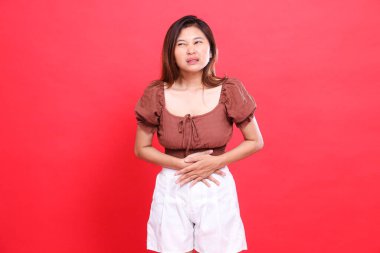Candid indonesia woman's expression in pain with stomach problems wearing a brown blouse and shorts on a red background. for health, lifestyle and advertising concepts clipart