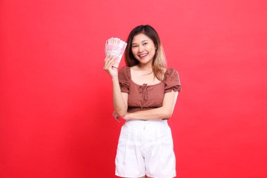 Neşeli Asyalı kadın, kırmızı arka planda kahverengi bir bluz giyerek, rupiah notaları tutarken kolları çaprazlanmış şekilde el kol hareketleri yapıyor. İşlem, teknoloji ve reklam kavramları için