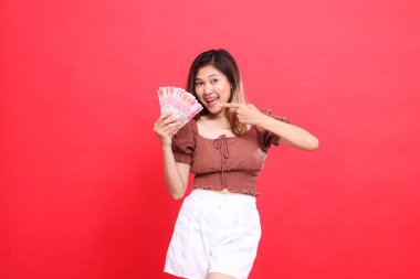 Kırmızı arka planda kahverengi bluz giyen ve elinde rupiah parası tutan neşeli, mutlu bir Asyalı kadının ifadesi. İşlem, teknoloji ve reklam kavramları için