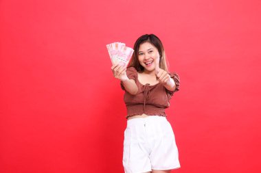 Neşeli, Asyalı genç bir kadının rupiah parasını ileri doğru tutması ve kırmızı arka planlı kahverengi bluz giyen bir baş parmak işareti vermesi. İşlem ve reklam için