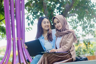 İki neşeli Asyalı kadın, kampüs parkında öğrencileri terfi ve yaşam tarzı kavramları için kucaklarken ellerinde kalemler, dizüstü bilgisayarlar, kitaplar ve panolarla birlikte çalışmayı tartışıyorlar.