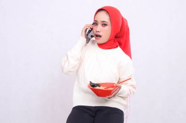 Başörtüsü takan Asyalı kadın cep telefonuyla arayınca şaşırdı ve içinde ramen (Çin yemeği) ve çubuk bulunan bir kase taşıyordu. Güzel Müslüman kadınlar reklam içeriği için kullanılır.