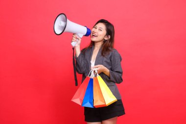 Endonezyalı iş kadını megafon hoparlörü üzerinden neşeli bir şekilde bağırıyor ve kırmızı arka planda gri bir ceket giyen bir alışveriş poşeti taşıyor. İşlem, yaşam tarzı ve iş kavramları için