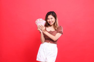 İki elinde rupiah para tutan neşeli Endonezyalı kadının ifadesi kırmızı arka planda kahverengi bir bluz giyiyor. İşlem, teknoloji ve reklam kavramları için