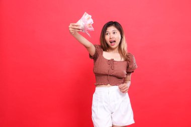 Şokta olan Endonezyalı genç bir kadının ifadesi. Elinde rupiah banknotları tutan ve elleri beline kadar uzanan kırmızı arka planda kahverengi bir bluz giyen. İşlem, teknoloji ve reklam kavramları için