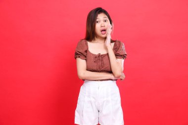 Fısıldaşma işareti olarak ellerini bir arada tutan, kahverengi bluz ve kırmızı arka planı olan şort giyen Asyalı bir kızın ifadesi. Moda, yaşam tarzı ve reklam için.