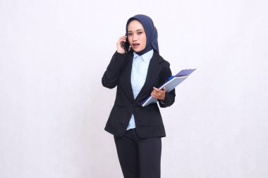 Özel tesettürlü Asyalı bir ofis kadını birinin telefon kamerasına şaşırmış ve yanında kalem, not defteri ve not defteri taşıyor. Mavi gömlekli güzel Müslüman kadın.