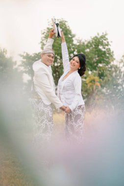 Bir çift genç Endonezya 'lı erkek ve kadın aşıklar kol kola, sırt sırta, modern Cava geleneksel giysileri içinde mutlu bir şekilde yürüyorlar, açık bir bahçede beyaz kadife gelinlik kebaya.