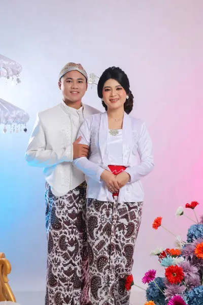 stock image A pair of Asian lovers smiling, the man's hand holding the woman's arm, wearing modern Javanese traditional clothing, a white velvet bridal kebaya, prewedding concept on a white studio background.