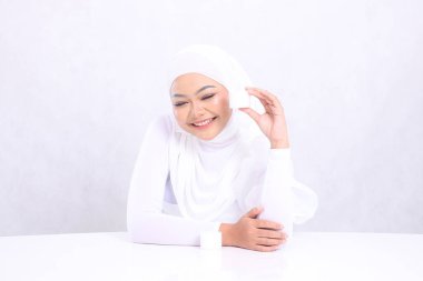Güzel yetişkin tesettürlü kız ifadesi beyaz arka planda günlük cilt bakımı için sol yüzünde kozmetik krem tutarak gülüyor. Yüz bakımı, mükemmel cilt.