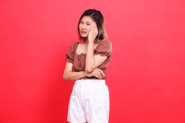 Endonezyalı bir kızın başı ağrılı, içten, kahverengi bluz ve kırmızı arka planlı şort giyen bir kızın ifadesi. Sağlık, bakım ve reklam konsepti için