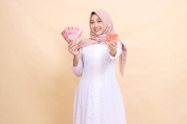 Başörtüsü takan bir kadın elinde rupiah parası ve kredi kartı tutarak neşeyle gülümsüyor. Yaşam tarzı, dini, iş ve finans kavramları