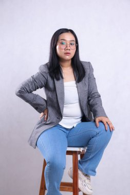 Güzel, genç bir ofis kadınının portresi. Zarif gözlükler takıyor, belini tutuyor, kalçaları gri kot pantolon giyiyor ve izole edilmiş beyaz arka planda oturuyor. Şirket, iş ve moda içeriği için