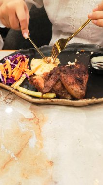 view of woman's hands enjoying dajaj mashwi, oven roasted chicken with tearing khubus (pita bread) with fork and knife accompanied by french fries, salata, chili sauce and vegetables on plate clipart