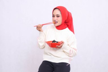 Başörtüsü takan Endonezyalı kadın kameraya bakar yemek çubuklarıyla eti ısırır ve içinde ramen olan bir kase (Çin yemeği) yer. Güzel Müslüman kadınlar mutfak, yemek ve reklam içerikleri için kullanılır.