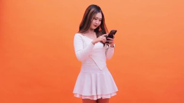 Asya kökenli zeki, neşeli, beyaz tişörtlü, parmakla işaret eden, cep telefonuyla kayan genç bir kadın, sade turuncu arka planda izole edilmiş büyük galibiyet haberini yeni öğrendi.