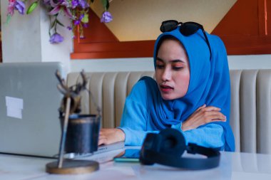 Genç Asyalı kız öğrenci omuz ağrısı nedeniyle acı içinde tesettür takıyor. Ev ödevi yapıyor. Kapalı kahve dükkanındaki kanepede oturuyor. Reklam içeriği, yaşam tarzı, çalışma için