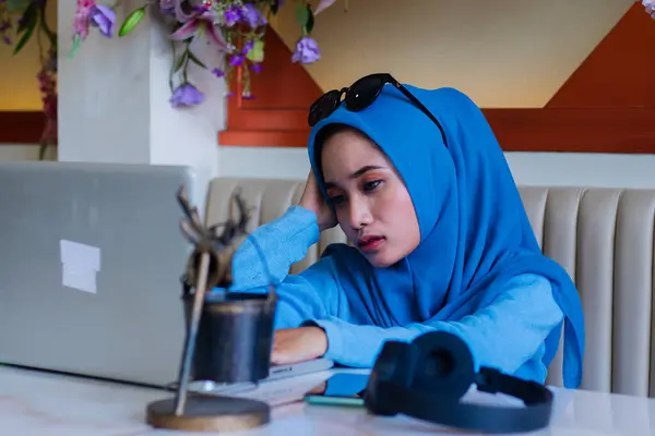 stock image young asian businesswoman wearing hijab suffering from headache because of thinking too much about her job sitting on sofa in indoor coffee shop. for advertising content, lifestyle, work