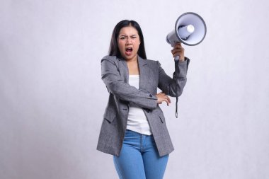 Beyaz arka planda izole edilmiş gri takım elbiseli, elinde hoparlörle kameraya kızgın bağıran genç ve güzel bir ofis kızı. moda, yaşam tarzı, reklamcılık, yayın, alışveriş içeriği