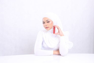 Güzel, zarif, genç bir tesettür genç bayan dudak makyajı görüntüsü kameranın eline bak ruj kabını tut ve masa izole edilmiş beyaz arka plana yapıştır. Güzellik eğitimi konsepti, dudak bakımı, ruj modeli