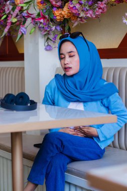 portrait of beautiful young asian girl wearing veil covering her eyes feeling unwell having stomach ache because of eating the wrong thing sitting on the sofa in coffee shop. for advertising,lifestyle clipart