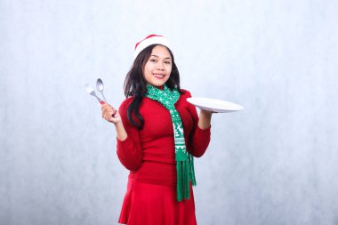 Endonezyalı kadının kırmızı kazak giyip Noel Baba şapkası ve atkısıyla mutlu noeller kutlaması yapması, kameraya gülümsemesi beyaz tabak taşıması ve beyaz arka planda izole edilmiş kaşık çatalı tutması.