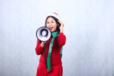 Neşeli yetişkin bir kadın iki elini de ileriye uzattı. Elinde hoparlör ve uzun saçlar vardı. Kırmızı Noel süveteri, Noel Baba şapkası ve atkısı vardı.