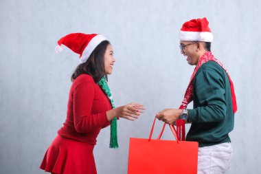 two young men wearing Christmas sweaters, Santa hats and scarves, cheerfully looking at their partner face while receiving Christmas gift paper bags, isolated on white background clipart