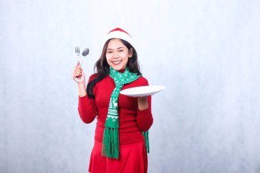 Noel Baba şapkası ve eşarbıyla kırmızı süveter giyen, beyaz tabak taşıyan, çatal tutan, mutlu bir yetişkin kadın. Mutlu noelleri ve beyaz arka planda izole edilmiş yeni yılı kutluyor.