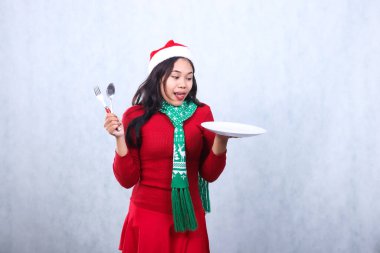 gesture young asian girl wearing red sweater with Santa hat and scarf, tempted to see food carry white plate and hold spoon fork, celebrate merry christmas and new year isolated on white background clipart