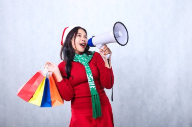 Neşeli, genç Asyalı kadın elini megafon hoparlörüne koydu ve renkli bir çanta taşıdı, kırmızı Noel süveteri giyip Noel Baba şapkası ve eşarbı taktı, beyaz arka planda izole edildi.