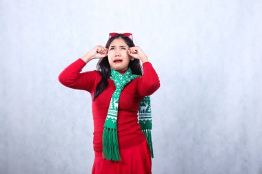 Noel Baba şapkası ve eşarpla kırmızı kazak giyen genç kız, mutlu noeller ve yeni yılı kutluyor, gözyaşlarını siliyor kafaya gözlük takıyor, beyaz arka planda soyutlanıyor.