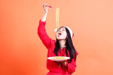 Genç bir kadının Noel elbisesi, Noel Baba şapkası, göz bağı üzerinden erişte koyması, kafasının üstünde erişte dolu el çatalı, erişte dolu beyaz tabak, izole edilmiş...