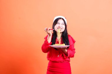 Makarna yiyen neşeli Asyalı kadın sola bakıyor, elinde çatalla erişte ve beyaz tabak taşıyor, kırmızı Noel bluzu giyiyor, Noel Baba şapkası takıyor, turuncu arka planda izole edilmiş.