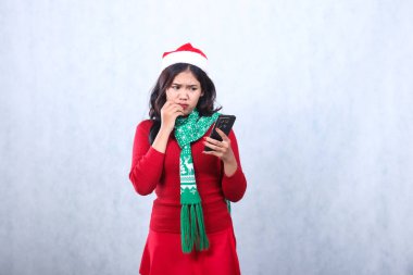 Charming young woman wearing red sweater with Santa hat and scarf, fidgeting to camera holding mobile phone while cupping lips, celebrating merry christmas and new year isolated on white background clipart