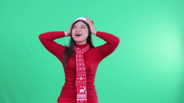 Noel süveteri giyen çekici Asyalı kadın, Noel Baba şapkası ve eşarbı, şen şakrak, yukarıdan gelen konfetiye bakan, yeşil arka planda izole edilmiş, yeşil ekran.