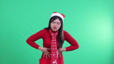 Genç Asyalı bir kadın midesi ağrırken, midesi ağrırken, adet öncesi karnı ağrırken, Noel süveteri, Noel Baba şapkası ve eşarp giyerken, yeşil perdede,.