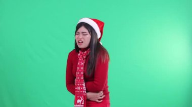 Endonezya 'lı genç bir kadın, yeşil arka planda, Noel süveteri, Noel Baba şapkası ve eşarbı giyerken, dayanılmaz bir acı ifadesi sergiliyor..