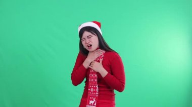 Endonezyalı kadın, yeşil ekranda Noel süveteri, Noel Baba şapkası ve eşarbı giyerken yutmakta zorlanıyor gibi görünürken boynunda acı ifadesi gösteriyor.