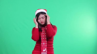 Noel süveteri giyen kız, Noel Baba şapkası ve eşarp takan ve baş ağrısı yüzünden acı çeken, kameraya bakan, iki eliyle başını tutarken, yeşil arka plan, yeşil ekran.