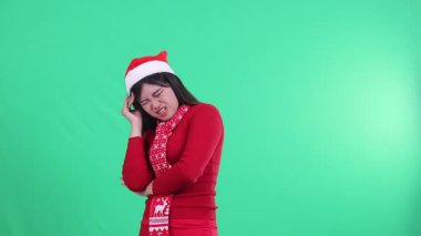 Kafasının sağ tarafı ağrıyan başını kollarında tutarak, Noel süveteri, Noel Baba şapkası ve eşarbı takarak yeşil bir arka planda izole edilmiş güzel bir kadının portresi.