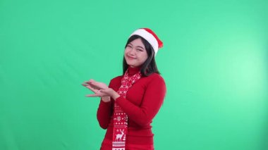 Asyalı genç bir kadının Noel süveteri, Noel Baba şapkası ve eşarbı giymesi, sağdaki el işaretinin gülümsemesi, yeşil arka planda izole edilmiş, yeşil ekran.