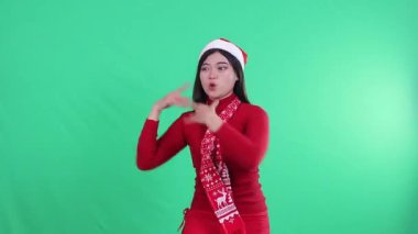 Asyalı genç bir kadının Noel süveteri, Noel Baba şapkası ve atkısı, sağda neşeli iki el, sağda işaret var, yeşil arka planda izole edilmiş, yeşil ekran.
