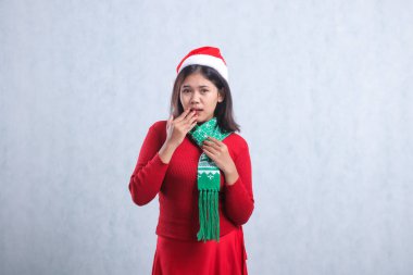 Endonezyalı genç bir kadının Noel süveteri, Noel Baba şapkası ve eşarbı giymesi, kameranın eline atkı tutmasından korkması, beyaz arka planda izole edilmiş bir şekilde.