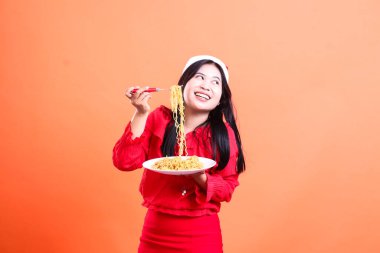 Kırmızı Noel bluzu giyen güzel bayanların jesti, Noel Baba şapkası, neşeli eğimli görünüm sol, beyaz tabaktan erişte dolu el çatalı, turuncu arka planda izole edilmiş.