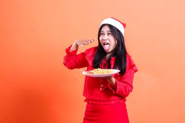Genç bir kadının Noel elbisesi, Noel Baba şapkası, dilinin kıvrımı baharatlı erişte yemek, ağızdan ağıza yellenmek, erişte dolu beyaz tabağı tutmak, izole edilmiş.