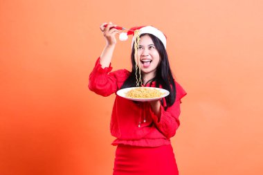 Neşeli genç Asyalı kadın erişteye bakıyor, elinde çatalla eriştelerle dolu beyaz tabak, kırmızı Noel bluzu, Noel Baba şapkası, turuncu arka planda izole edilmiş.